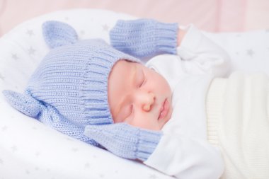 Sleeping little baby wearing knitted blue hat with ears and mittens lying in beautiful cradle. Security and childcare concept clipart