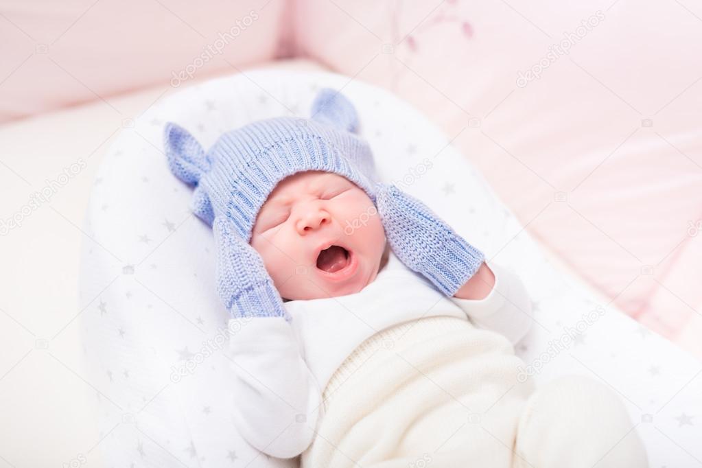 Yawning little baby wearing knitted blue hat with ears and mittens lying in beautiful cradle. Security and childcare concept