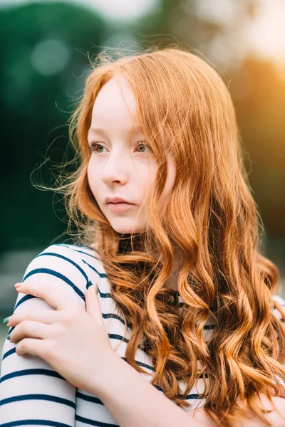 Una hermosa adolescente con el pelo largo y rizado de jengibre y ojos verdes abrazándose en el parque de verano. Retrato al aire libre de la atractiva joven pelirroja en la noche al atardecer — Foto de Stock