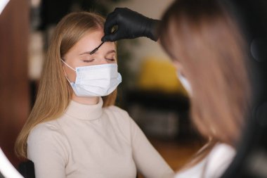 Koruyucu maskeli ok ve makyaj ustası, daha önce bir güzellik salonunda kınalı kaşlarla boyanmış forseps ile çıkartmak için şekil verir. Tıbbi maskeli güzel sarı saç modeli.