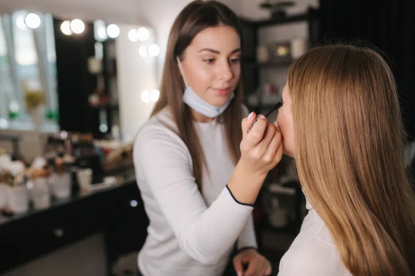 Artista de maquiagem feminina trabalhando no salão de beleza durante a quarentena. Maquiagem artista em máscara médica no queixo. Modelo de cabelo loiro bonito — Fotografia de Stock