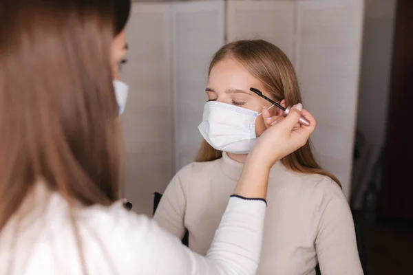 Karantina sırasında güzellik salonunda çalışan bir kadın makyöz. Tıbbi maskeli bir makyöz. Koruyucu maskeli güzel sarı saç modeli — Stok fotoğraf