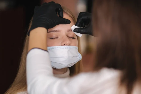 Brow and makeup master in protective mask gives shape to pull out with forceps previously painted with henna eyebrows in a beauty salon. Beautiful blond hair model in medical mask