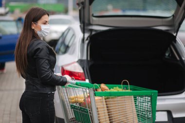 Çekici genç bir kadın market alışverişini tramvaydan araba kamyonuna taşıyor. Enfeksiyonu önlemek için sosyal güvenlik mesafesi, yüz maskesi, tek kullanımlık eldivenler. Yiyecek alışverişi sırasında
