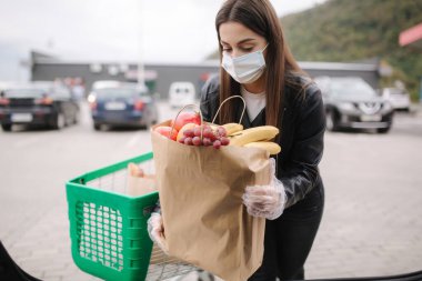 Süpermarketten alışveriş yaptıktan sonra bagaja maske takan tıbbi maskeli genç bir kadının bagaj görüntüsü. Quqrantine market nakliyesi. Büyükbabalar için taze meyve. Eko ustası kadın paket