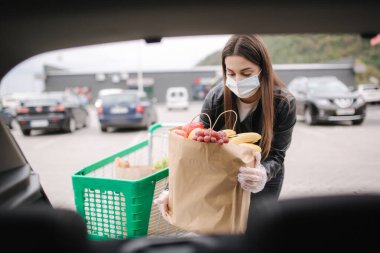 Süpermarketten alışveriş yaptıktan sonra bagaja maske takan tıbbi maskeli genç bir kadının bagaj görüntüsü. Quqrantine market nakliyesi. Büyükbabalar için taze meyve. Eko ustası kadın paket