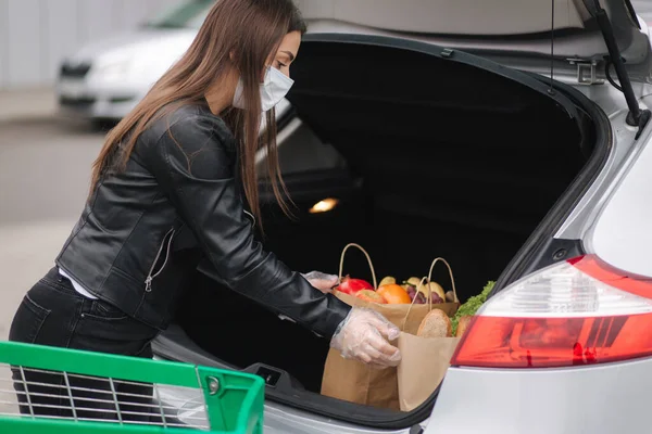 一个过程，年轻迷人的女人把食品从超市从推车到汽车卡车。社会距离保护，面具，一次性手套，以防止感染。食物 — 图库照片