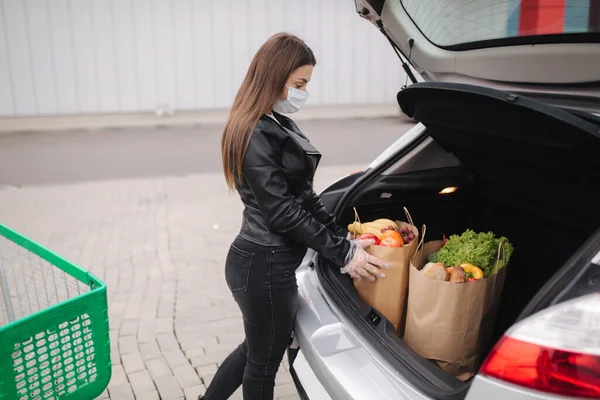 一个过程，年轻迷人的女人把食品从超市从推车到汽车卡车。社会距离保护，面具，一次性手套，以防止感染。食物 — 图库照片