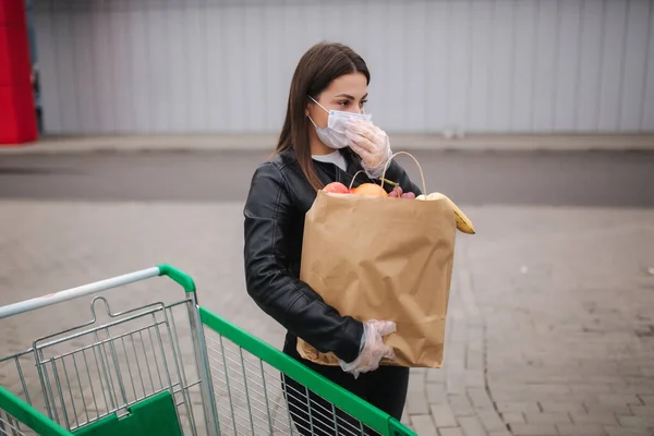 Mujer joven con máscara protectora y guantes desechables lleva un paquete de artesanía con comestibles a su coche en frente del supermercado — Foto de Stock