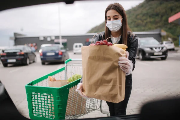 Над багажником вид молодой женщины в медицинской маске в масках погрузки сумки в багажник после покупки супермаркета. Доставка продуктов питания Quqrantine. Свежие фрукты для бабушки и дедушки. Женщина с экологическим ремеслом пакет — стоковое фото
