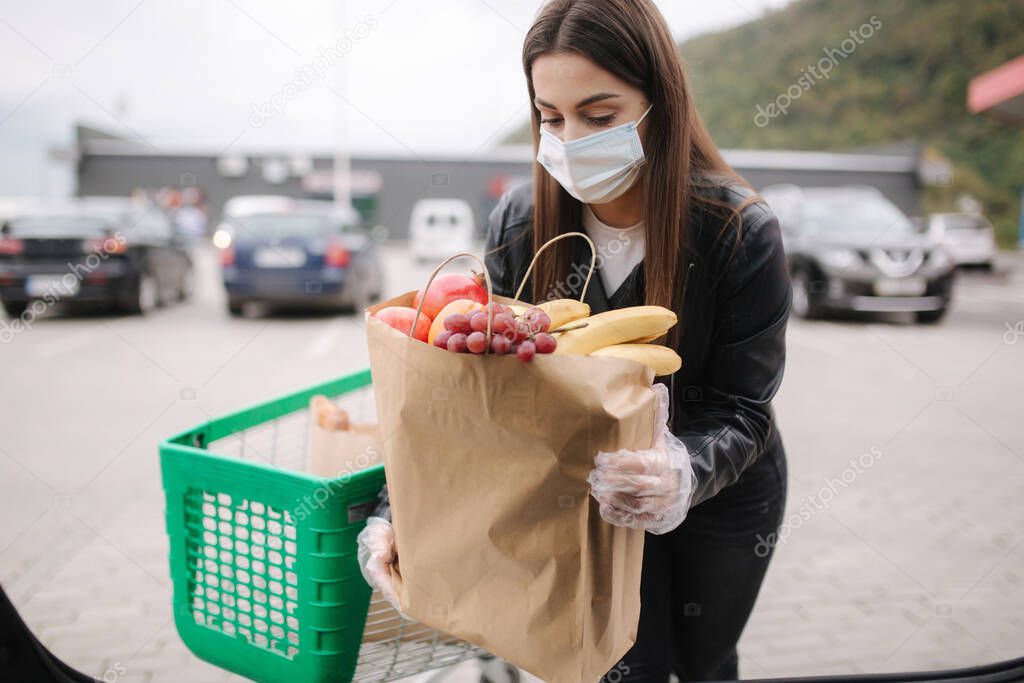 Over trunk view of young woman in medical mask in masks loading bags in trunk after supermarket shopping. Quqrantine grocery shipping. Fresh fruits for grandparents. Female with eco craft paket