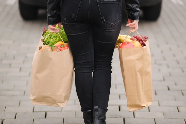 Back view of woman in black clothes carries a eco package full of griceries to the car. Fresh vegetables and fruits. Midle selection
