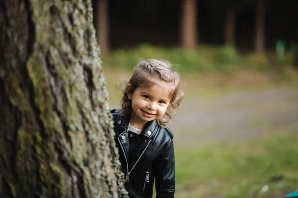 Маленькая девочка смотрит с дерева в лесу. Милая маленькая девочка с красивыми косичками в кожаной куртке — стоковое фото