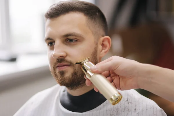 Peluquería profesional utiliza un cortador de pelo para flecos barba para un hombre guapo en la barbería — Foto de Stock