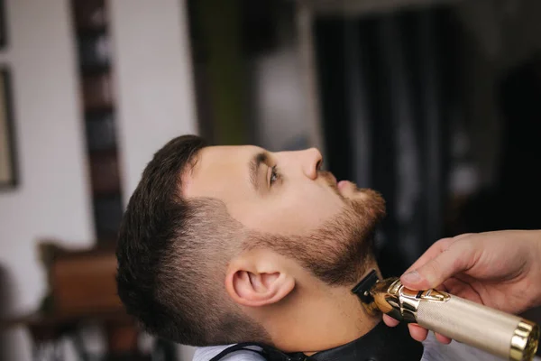 Peluquería profesional utiliza un cortador de pelo para la barba de flecos para un hombre guapo en la barbería. Vista lateral — Foto de Stock