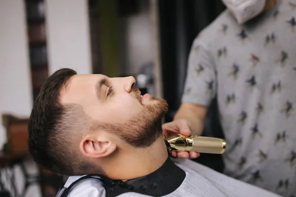 Peluquería profesional utiliza un cortador de pelo para la barba de flecos para un hombre guapo en la barbería. Vista lateral — Foto de Stock