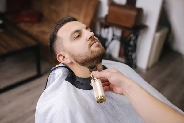 Peluquería profesional utiliza un cortador de pelo para la barba de flecos para un hombre guapo en la barbería. Vista en primera persona — Foto de Stock