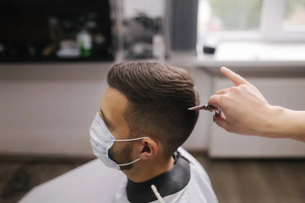 Peluquero cortándole el pelo a un guapo barbudo con máscara facial. Máscara protectora en la cara del cliente. La peluquera también lleva una máscara. concepto de corte de pelo cuarentena. — Foto de Stock