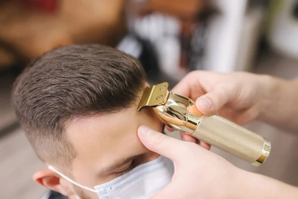 Peluquería profesional utiliza un cortador de pelo para flecos cabello para un hombre portador guapo. El cliente con máscara facial protege del coronavirus. La peluquera también lleva una máscara — Foto de Stock