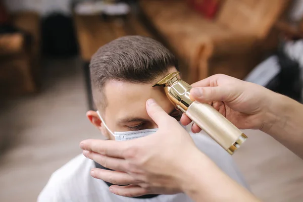 Professionele kapper maakt gebruik van een tondeuse voor fringing haar voor een knappe drager man. Cliënt met gezichtsmasker beschermt tegen het coronavirus. De kapper draagt ook een masker — Stockfoto