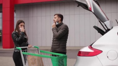 Karantina sırasında süpermarket alışverişinden sonra bagaja çanta yükleyen maskeli genç bir çift. Karısıyla birlikte açık hava otoparkında bir adam. Taze yiyecekle birlikte çevre paketi