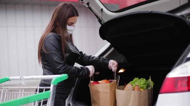 Mladá žena v ochranné masce a jednorázových rukavicích nese do auta před supermarketem řemeslný balíček s potravinami — Stock video