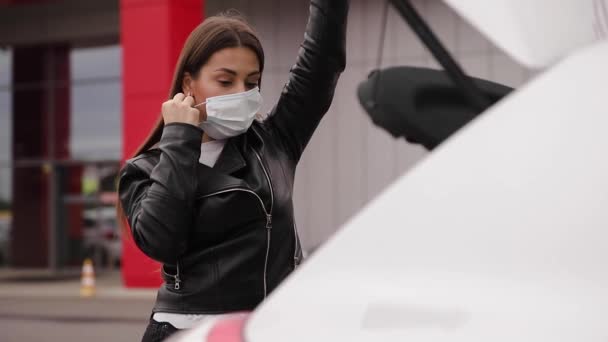 Young stylish woman in protective mask with shopping cart full of fresh food during quarantine, packing products into the car on the outdoor parking. Woman close car trunk — Stock Video