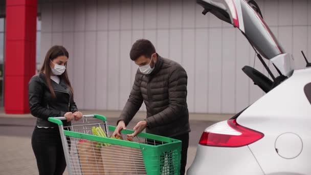 Mladý pár v maskách nakládá tašky v kufru po nakupování v supermarketu během karantény. Muž s její ženou na venkovním parkovišti. Eko balíček s čerstvým jídlem — Stock video