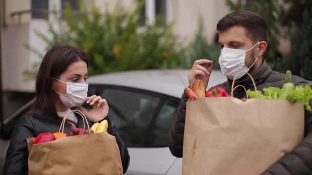 Pasangan muda bertopeng memegang paket eko dengan makanan segar setelah belanja supermarket selama karantina. Pria dengan istrinya di rumah melepas topeng — Stok Video