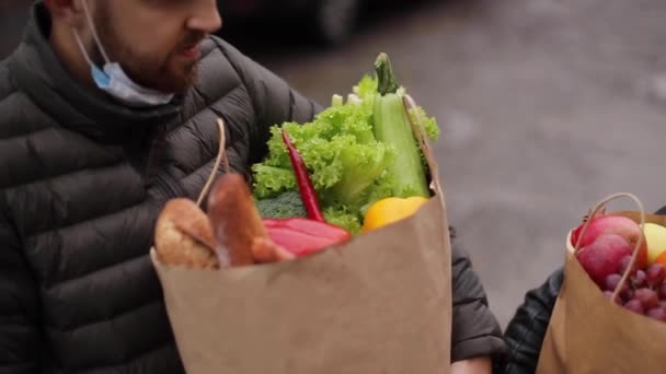 Крупным планом молодая пара в масках держит эко-пакет со свежей едой после покупок в супермаркете во время карантина. Мужчина и женщина в защитной маске на подбородке — стоковое видео
