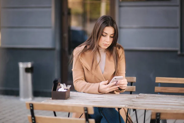 Aantrekkelijke jonge vrouw zit op terrasje in cafe en het gebruik van de telefoon. Mooie brunette zit buiten en chatten — Stockfoto