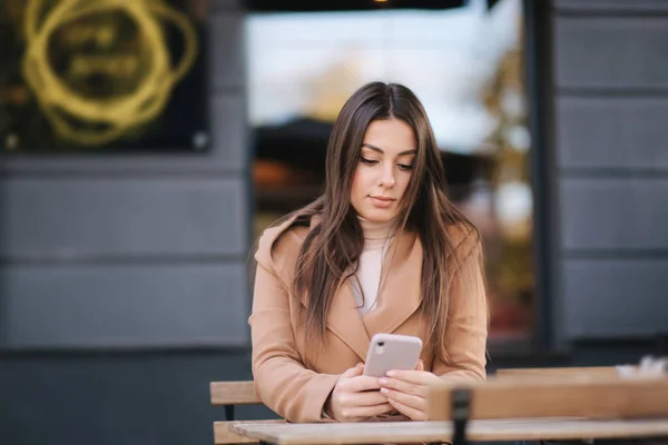 Aantrekkelijke jonge vrouw zit op terrasje in cafe en het gebruik van de telefoon. Mooie brunette zit buiten en chatten — Stockfoto