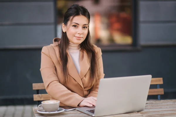 Jonge zakenvrouw zittend op terras door cafe met laptop. Een vrouw die koffie drinkt en buiten werkt. Vrouw gebruik telefoon en notebook — Stockfoto