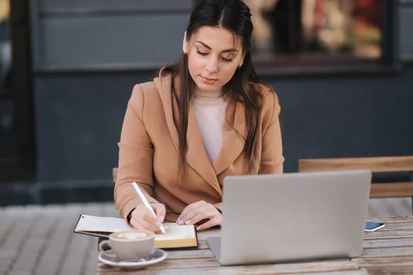 Ελκυστική νεαρή επιχειρηματίας που γράφει κάτι στο σημειωματάριο. Γυναικείος καφές και εργασία σε εξωτερικούς χώρους χρησιμοποιώντας φορητό υπολογιστή — Φωτογραφία Αρχείου