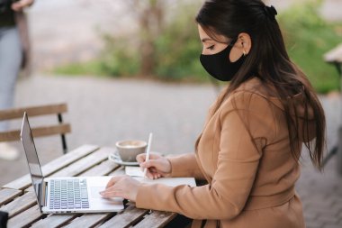 Kafede terasta oturan siyah maskeli genç bir kadın. Karantinada çalışan bir iş kadını. Yan görüş. Covid 19