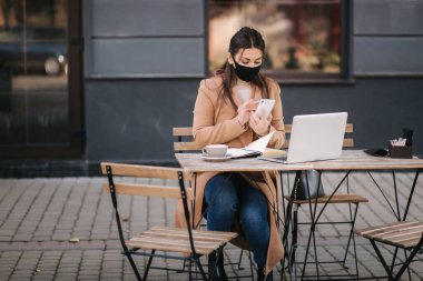 Karantina sırasında kafede terasta oturan siyah maskeli genç bir kadın. Karantinada çalışan bir iş kadını. Kadın telefonda konuşuyor. Covid 19