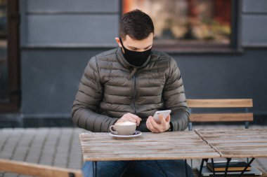 Kafenin yanındaki terasta telefon kullanarak siyah maskeli yakışıklı bir adam. Karantina sırasında kafede bir adam var. Covid 19