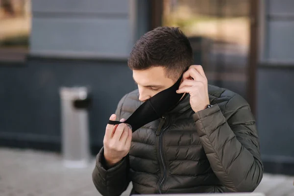 Handsome man putting on black protective mask on terrace by the cafe. Man outdoors during quarantine. Covid-19 — Stock Photo, Image