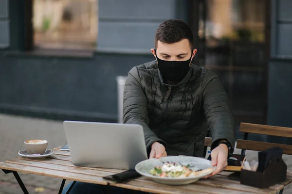 Handsome man in face mask sitting outdoors in cafe and working on laptop. Man take salad. Quarantine. Covid-19