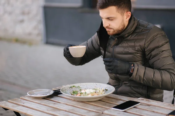 Man in black gloves hold cutlery. Quarantine Cafe concept. Food outdoors in protective gloves. Fresh vegan salad with shrimps. Man pulled off protective mask for drinking cappuccino