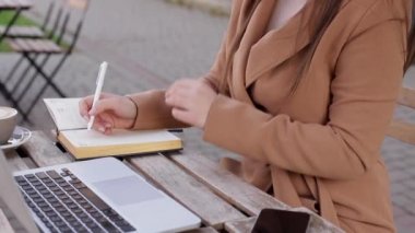 Dışarıdaki kafede çalışma süreçlerini kapatın. Deftere bir şeyler yazan bir kadın. Masada cappuccino laptop ve takvim