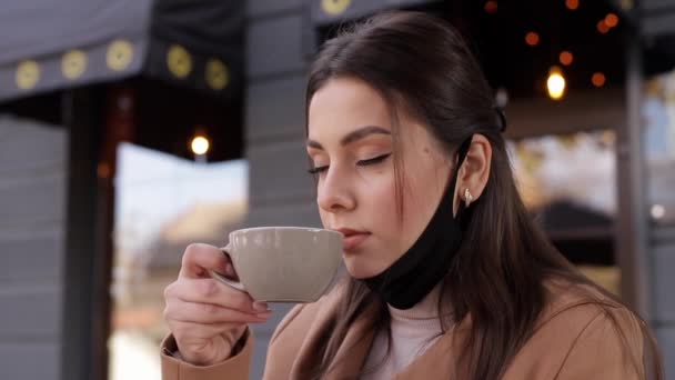 Proceso de hermosa mujer se quita la máscara protectora para beber café. Mujer sentada al aire libre en la terraza y beber café. Cuarentena, autodistanciamiento. Covid-19 — Vídeos de Stock