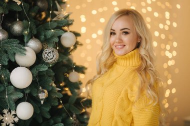 Evdeki büyük Noel ağacının yanında çekici genç bir anne. Sarı saçlı, süveter giyen kadın gülümsedi.