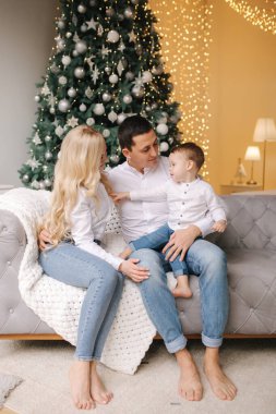 Baba, anne ve oğlunun portresi evde, Noel ağacının yanında yerde oturuyor, hepsi gülümsüyor. Denim ve beyaz gömlekler. Noel ailesi. Mutluluk