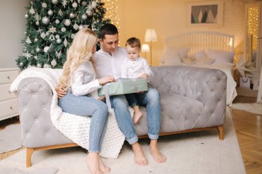 Baba, anne ve oğlunun portresi evde, Noel ağacının yanında yerde oturuyor, hepsi gülümsüyor. Denim ve beyaz gömlekler. Noel ailesi. Mutluluk