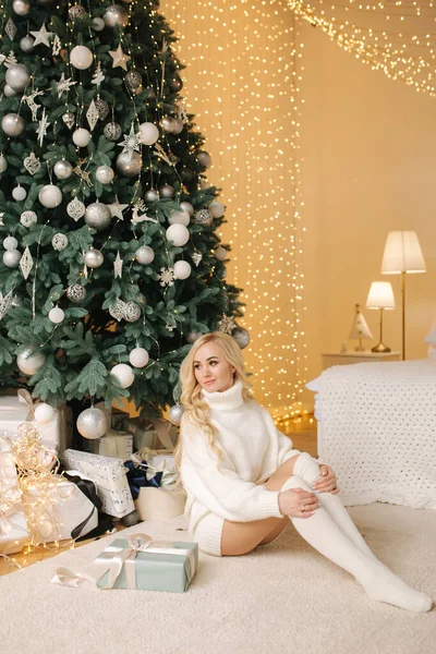 Vacker blond hår kvinna i vit luvtröja och ledde värmare sitter framför stora julgran. Sexig kvinna sitter på golvet hemma på semester — Stockfoto