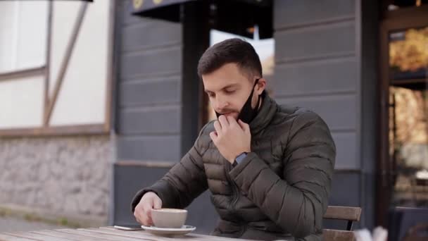 Mann zog Gesichtsmaske für trinkenden Cappuccino auf Terrasse ab Mann in Café im Freien während Quarantäne. Covid-19 — Stockvideo