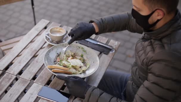 İnsanoğlu, yemek yemek için koruyucu maskeyi çıkardı. Siyah eldivenli adam çatal bıçak takımı tutuyor. Karantina Kafe konsepti. Koruyucu eldivenlerle dışarıda yemek. Karidesli taze vejetaryen salatası. Yan görünüm — Stok video