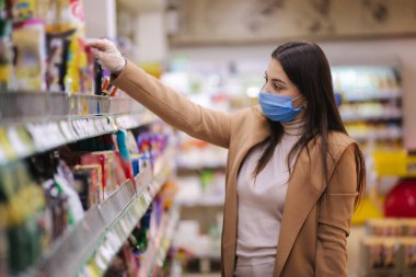 Koruyucu maskeli ve eldivenli bir kadının süpermarkette dikilirken mallara fiyat etiketi okuduğu yatay bir fotoğraf. Güzel genç kız markette alışveriş yaparken yılan seçiyor.