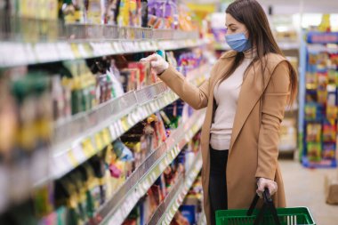 Koruyucu maskeli ve eldivenli bir kadının süpermarkette dikilirken mallara fiyat etiketi okuduğu yatay bir fotoğraf. Güzel genç kız markette alışveriş yaparken yılan seçiyor.
